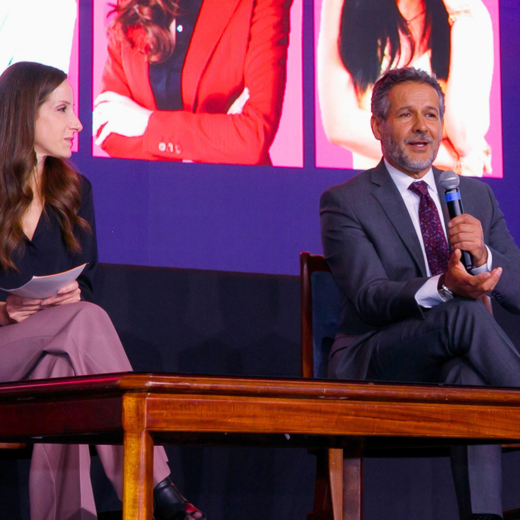 Two panelists sit on a stage: a woman on the left holds papers while a man in a suit speaks into a microphone on the right.